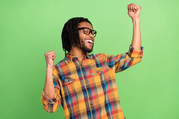 Photo of dreamy lucky dark skin guy dressed checkered shirt glasses dancing rising fists smiling isolated green color background — Stock Photo, Image