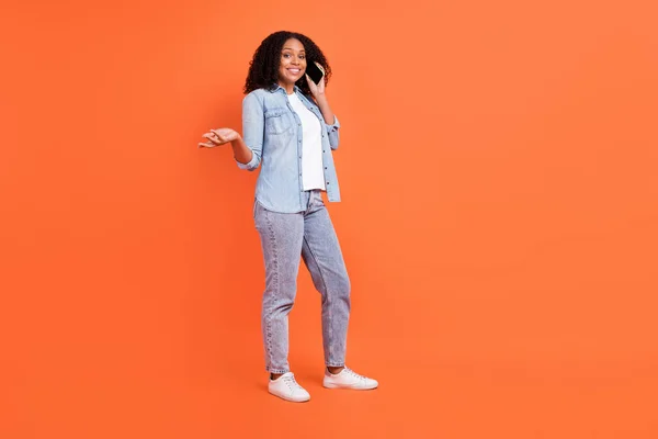 Helkroppsfoto av osäker ung brunett dam prata telefon bära jeans skjorta skor isolerad på orange bakgrund — Stockfoto