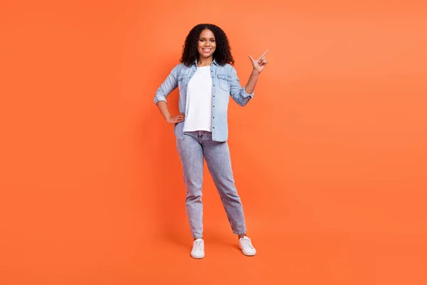 Full size foto van mooie jonge brunette dame punt lege ruimte dragen jeans shirt schoenen geïsoleerd op oranje achtergrond — Stockfoto