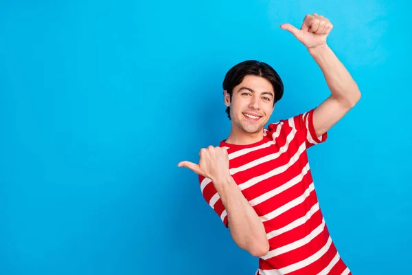 Фото очаровательного молодого парня, одетого в красную футболку, указывающую большими пальцами назад пустое пространство, улыбающегося изолированного синего цвета фона — стоковое фото