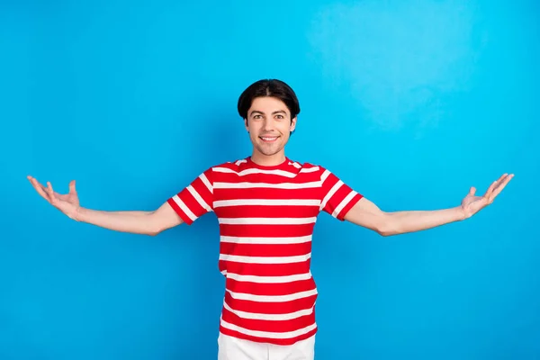 Foto van lieve schattige jonge man dragen gestreepte t-shirt glimlachen vergelijken armen lege ruimte geïsoleerde blauwe kleur achtergrond — Stockfoto