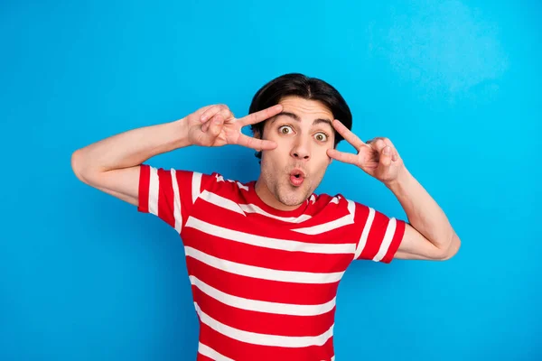 Foto van schattige geschokte jonge man dragen gestreepte t-shirt tonen v-tekens cover ogen geïsoleerde blauwe kleur achtergrond — Stockfoto