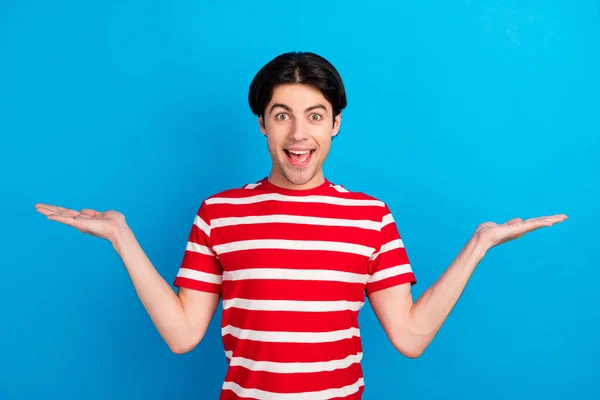 Foto de amigable joven emocionado vestido camiseta roja sonriendo mostrando las escamas de brazos espacio vacío aislado color azul fondo — Foto de Stock