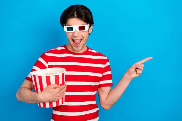 Foto van geschokte schattige jonge man gekleed rood t-shirt 3D-brillen eten pop maïs wijzend lege ruimte geïsoleerde blauwe kleur achtergrond — Stockfoto