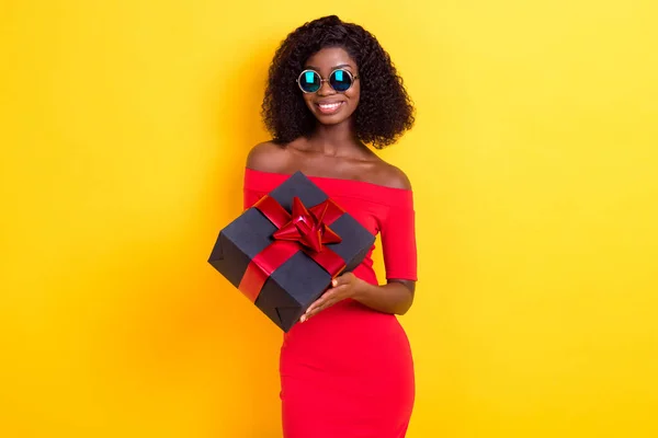 Foto de jovem encantador sorrindo bom humor afro menina em óculos de sol realizar presente de aniversário isolado no fundo de cor amarela — Fotografia de Stock