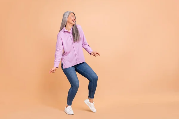 Фотографія повного розміру красивого білого зачіски у віці леді танцює носить фіолетові сорочки джинси кросівки ізольовані на бежевому фоні — стокове фото