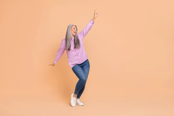 Фотографія повної довжини красивого сірого волосся стара леді танцює носить фіолетові сорочки джинси взуття ізольовані на бежевому фоні — стокове фото