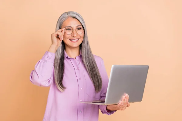 Foto de bonito peinado blanco señora anciana celebrar portátil usar gafas reflectante pantalla púrpura camisa aislada sobre fondo beige —  Fotos de Stock