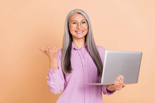 Zdjęcie optymistyczne szare włosy starsza pani trzymać laptop indeks promocyjne nosić okulary fioletowy koszula izolowana na beżowym tle — Zdjęcie stockowe