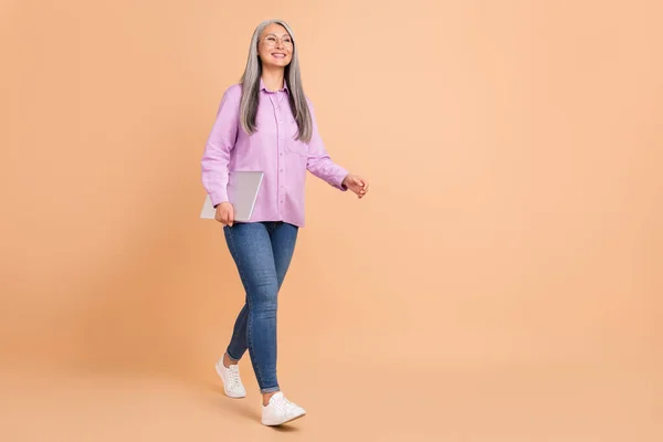 Foto de tamaño completo de buen peinado blanco anciana celebrar portátil ir mirada gafas camisa jeans zapatillas aisladas sobre fondo beige —  Fotos de Stock