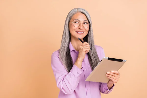 Foto de bonito penteado cinza mais velho senhora segurar tablet olhar espaço vazio desgaste óculos camisa violeta isolado no fundo cor bege — Fotografia de Stock