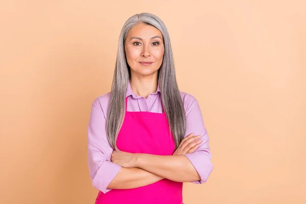 Foto van streng grijs kapsel oud dame gekruiste armen dragen paars shirt roze schort geïsoleerd op beige kleur achtergrond — Stockfoto