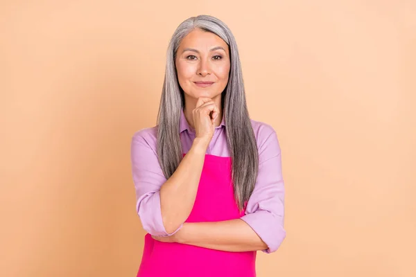Foto de la señora mayor del peinado gris inteligente piensa usar camisa púrpura delantal rosa aislado sobre fondo de color beige — Foto de Stock