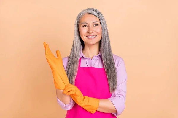 Portret van aantrekkelijke vrolijke grijs-harige vrouw zetten handschoenen op dagelijks opruimen geïsoleerd over beige pastel kleur achtergrond — Stockfoto
