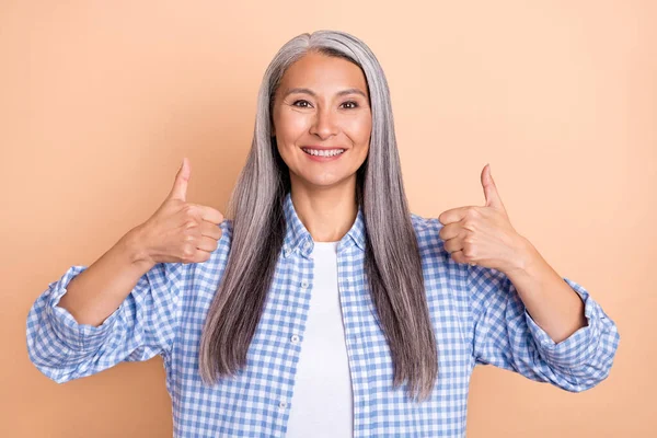 Фото чарівної милої леді пенсіонерки, одягненої в картату сорочку, що показує два великі пальці вгору посміхаючись ізольований бежевий колір фону — стокове фото