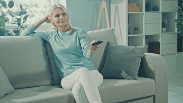 Lystig, avslappet, drømmende dame sitte komfortabel med sofaen og holde fjernkontroll på tv – stockvideo