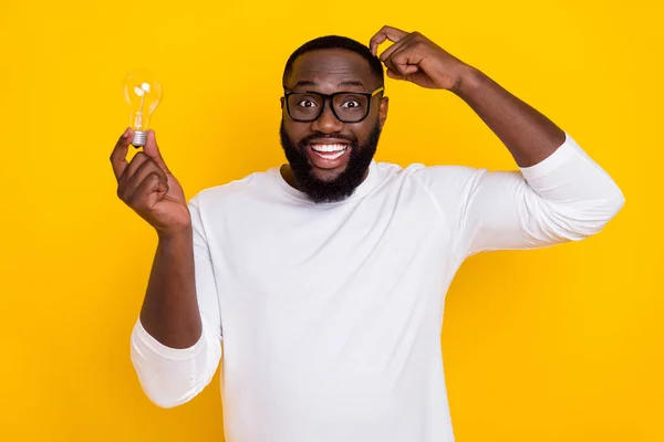 Photo of funky positive handsome guy hold light bulb scratching head toothy smile isolated on yellow color background — Stock Photo, Image