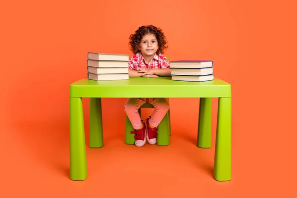 Foto de linda chica de la escuela divertida vestida a cuadros ropa sentada mesa lectura libros pila aislado color naranja fondo —  Fotos de Stock