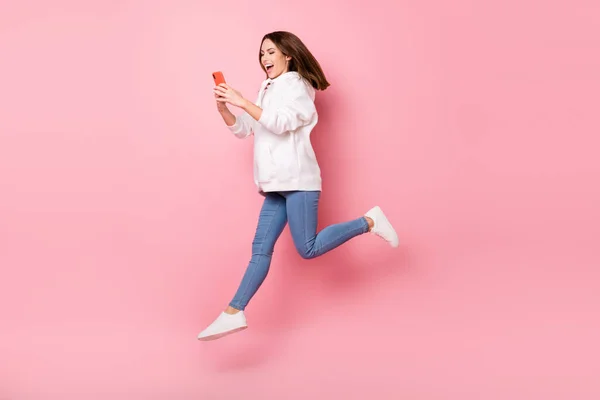Foto av bedårande upphetsad ung dam bära vit hoodie leende hoppa hög typing modern enhet isolerad rosa färg bakgrund — Stockfoto