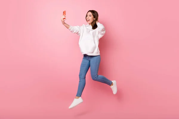 Photo drôle jeune femme mignonne habillée en sweat-shirt blanc tache selfie sautant haute sourire isolé fond de couleur rose — Photo