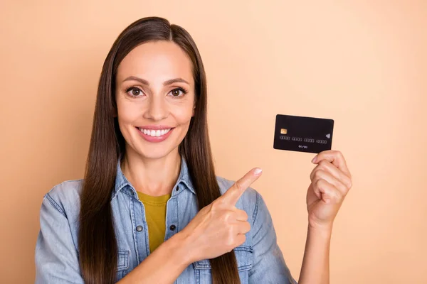 Фото веселой женщины, рекламирующей кредитную карту, указывающей пальцем на ношение джинсовой рубашки, изолированной на бежевом фоне — стоковое фото