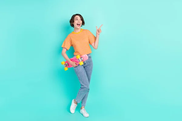 Photo of funky excited woman dressed striped t-shirt smiling holding skateboard pointing empty space isolated teal color background — Stock Photo, Image