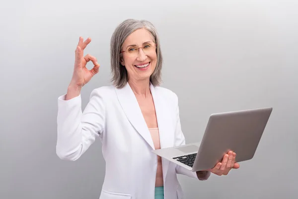 Photo of cool white hairdo old bossy lady hold laptop show okey wear spectacles jacket isolated on grey color background — Stock Photo, Image