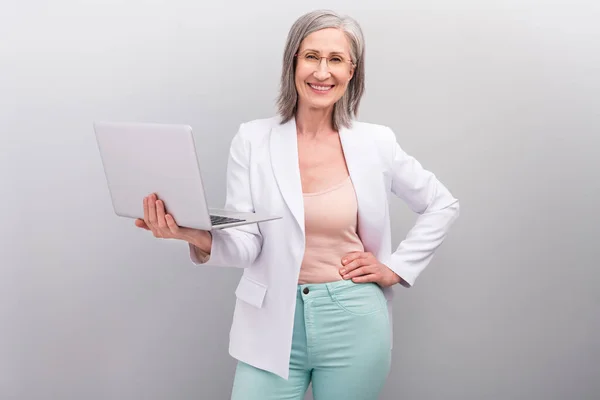 Patronluk taslayan beyaz saçlı yaşlı öğretmen hanımın fotoğrafı. Dizüstü bilgisayara bak. Gri arka planda izole edilmiş gözlük ceketi giy. — Stok fotoğraf