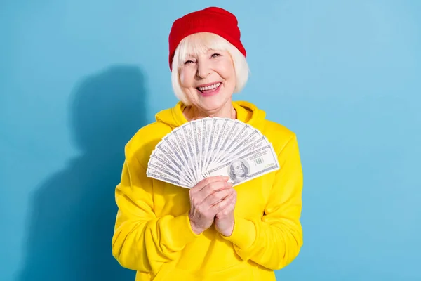 Foto di fortunato divertente età donna vestito giallo felpa rosso copricapo tenendo ventilatore usd centinaia isolato colore blu sfondo — Foto Stock
