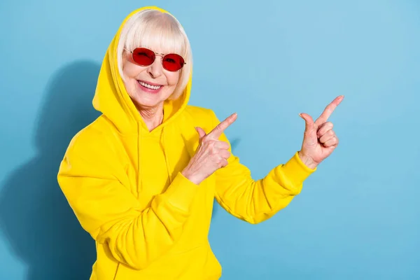 Foto de doce bonita velha mulher apontar dedos espaço vazio anúncio usar capuz de vidro solar isolado no fundo de cor azul — Fotografia de Stock