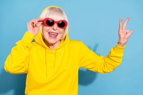 Фото веселой милой счастливой пожилой женщины носить солнцезащитные очки капюшон сделать V-знак изолированы на синем фоне цвета — стоковое фото