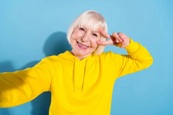 Fotografie veselý pěkný šťastný stará žena, aby selfie držet kamera show V-sign izolované na modrém pozadí — Stock fotografie