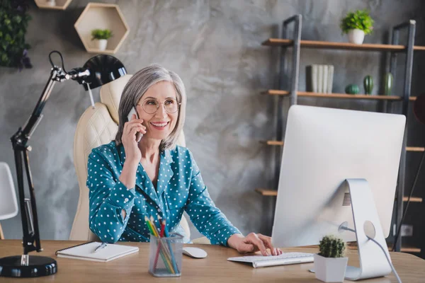 Oldukça olumlu bir fotoğraf. Güzel, mutlu, yaşlı, gri saçlı bir kadın. Masaya oturmuş, iş yerinin içinde telefonla konuşuyor. — Stok fotoğraf