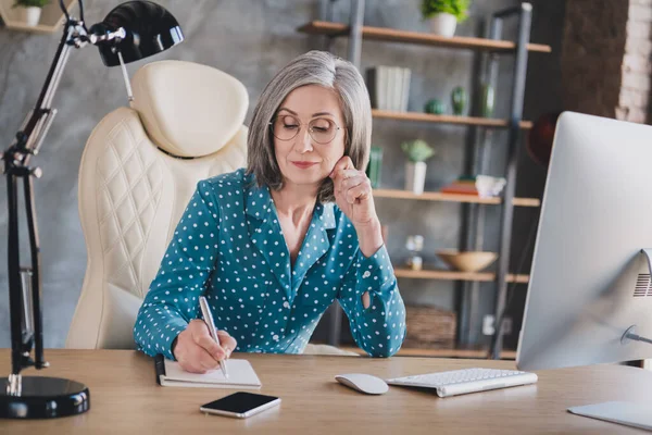 Foto allvarlig lugn gammal ganska gråhårig kvinna bära glasögon skriva anteckning manager titta inomhus på arbetsplatsen arbetsstation — Stockfoto