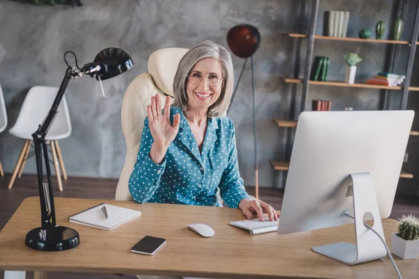 Fotografie atraktivní šťastný starý žena vlna ahoj sedět stůl kancelář uvnitř domu pracoviště — Stock fotografie