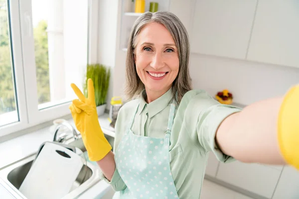 Autorretrato de atractiva mujer de pelo gris alegre haciendo platos aseado limpiar mostrando v-signo en casa blanco claro interior — Foto de Stock