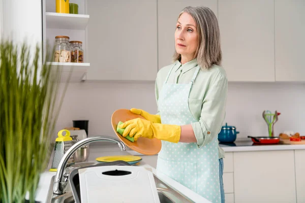 Çekici, gri saçlı bir kadının portresi. Bulaşıkları yıkıyor, evi temizliyor. Açık beyaz bir ev. — Stok fotoğraf