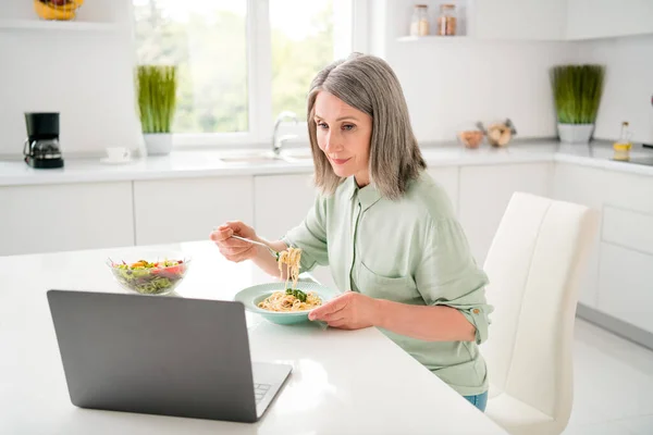 Πορτρέτο της ελκυστική εστιασμένη γκρίζα μαλλιά γυναίκα τρώει σπιτικό δείπνο βλέποντας βίντεο στο σπίτι φως λευκό εσωτερικό — Φωτογραφία Αρχείου