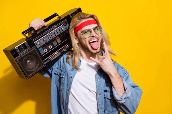 Foto de cara rude encantador vestido camisa jeans sorrindo mostrando língua sinal de rocha segurando caixa de boom isolado cor amarela fundo — Fotografia de Stock