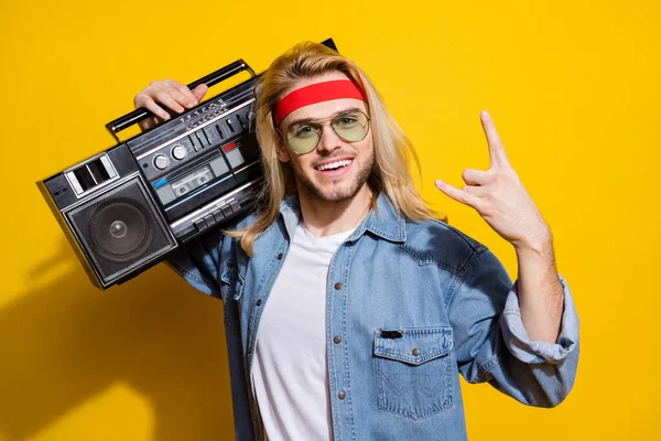 Zdjęcie cute cool gentleman nosić dżinsy koszula ciemne okulary słuchanie boombox pokazując hard rock znak odizolowany żółty kolor tło — Zdjęcie stockowe