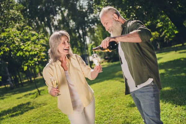 Foto de positivo aposentado partido casal homem segurar garrafa derramar copo de champanhe desgaste casual roupa natureza parque jardim fora — Fotografia de Stock