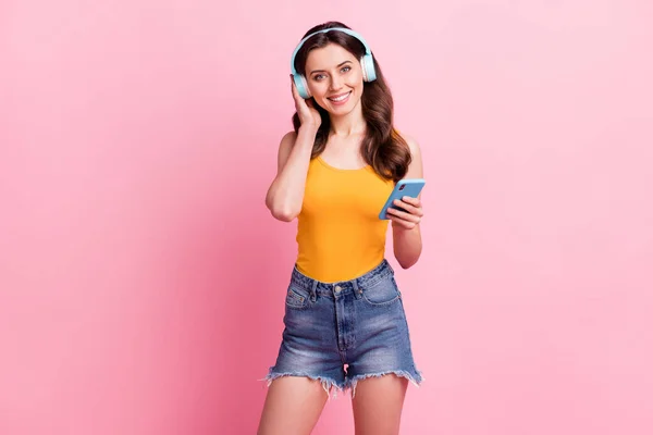 Foto de brillante encantadora joven vestida con camiseta amarilla que sostiene el dispositivo moderno escuchando canciones aisladas de color rosa de fondo — Foto de Stock
