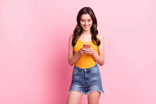 Foto de brilhante muito jovem senhora vestida amarelo tanque-top digitando dispositivo moderno sorrindo espaço vazio isolado cor de fundo rosa — Fotografia de Stock