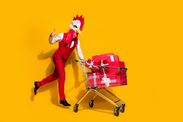 Foto de chico inusual celebrar carro de mercado comprar regalos levantar el pulgar hacia arriba usar máscara de polla traje rojo aislado color amarillo fondo — Foto de Stock