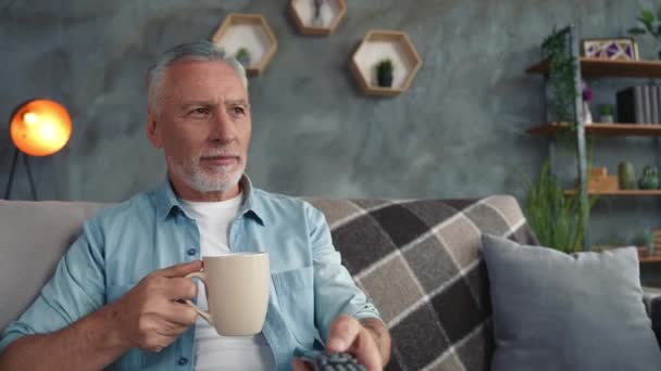 Mediana edad hombre sentarse sofá chill ver comedia bebida latte en moderno salón — Vídeos de Stock