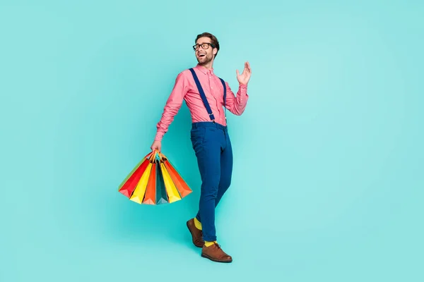 Foto lateral de perfil de tamaño completo del joven sonrisa feliz ir caminar mano ola hola bolsas de compras aisladas sobre fondo de color turquesa —  Fotos de Stock