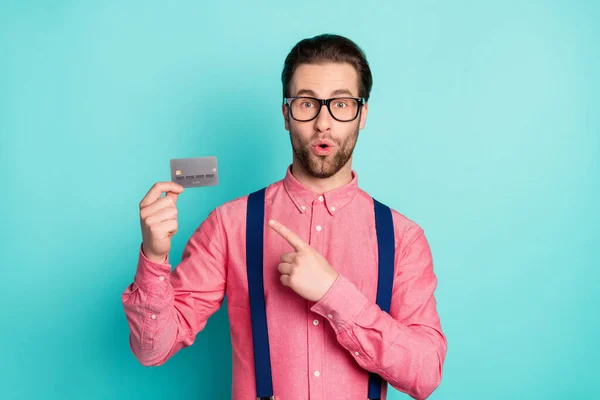 照片给人印象深刻的可爱小伙子戴着粉色衬衫眼镜指着手指信用卡孤立的绿松石色背景 — 图库照片