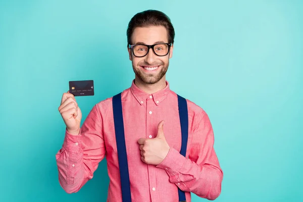 Foto van funky mooie heer dragen roze shirt bril met credit card tonen duim omhoog glimlachen geïsoleerde teal kleur achtergrond — Stockfoto