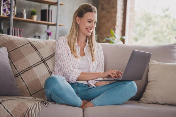 Полная длина фото умная зрелая блондинка сидеть на диване работа ноутбук носить рубашку джинсы дома в комнате в одиночку — стоковое фото