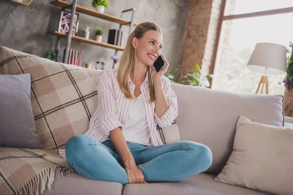 Полноразмерное фото очаровательной зрелой блондинки, сидящей на диване, разговаривающей по телефону, одетой в джинсы в белой рубашке дома в карантине — стоковое фото